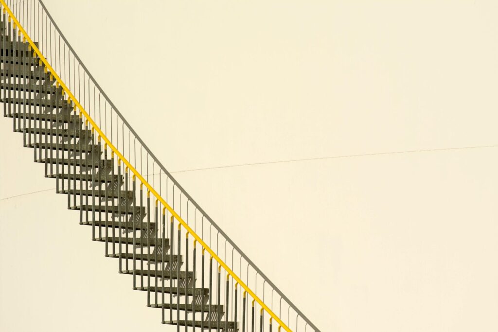 A metal staircase with yellow railing against a minimalistic beige background, showcasing industrial design.