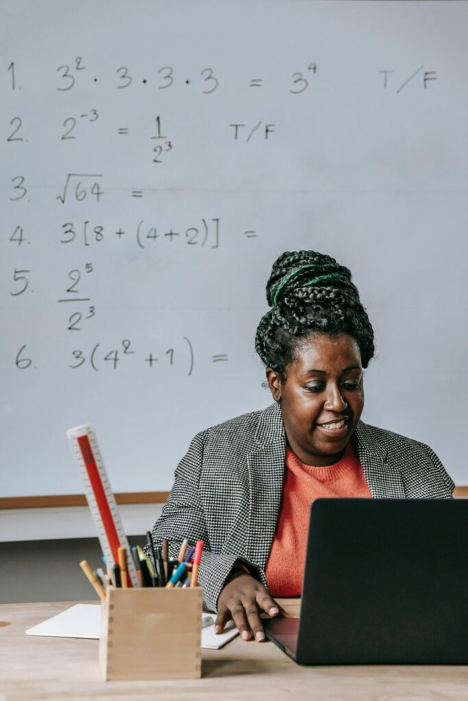 African American female teacher in formal clothes sitting at table with netbook and office supplies and looking at screen