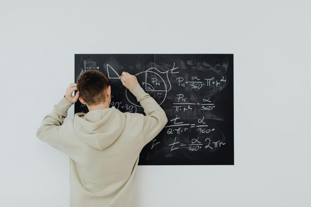 Teenager in hoodie writing math formulas on a blackboard indoors, showcasing problem-solving skills.