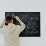 Teenager in hoodie writing math formulas on a blackboard indoors, showcasing problem-solving skills.