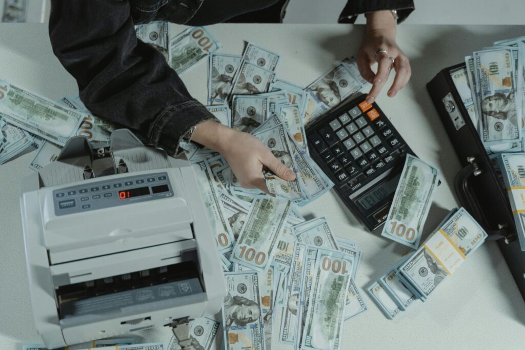 Hands counting 100 dollar bills using a calculator and money counter on a table.