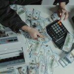 Hands counting 100 dollar bills using a calculator and money counter on a table.