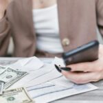 Close-up of a woman using a smartphone, holding cash, and managing finances indoors.