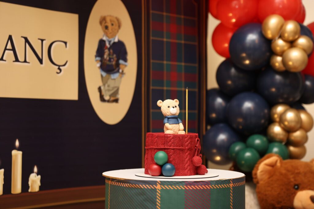 Teddy bear cake topper with vibrant balloons and plaid backdrop at a festive celebration.