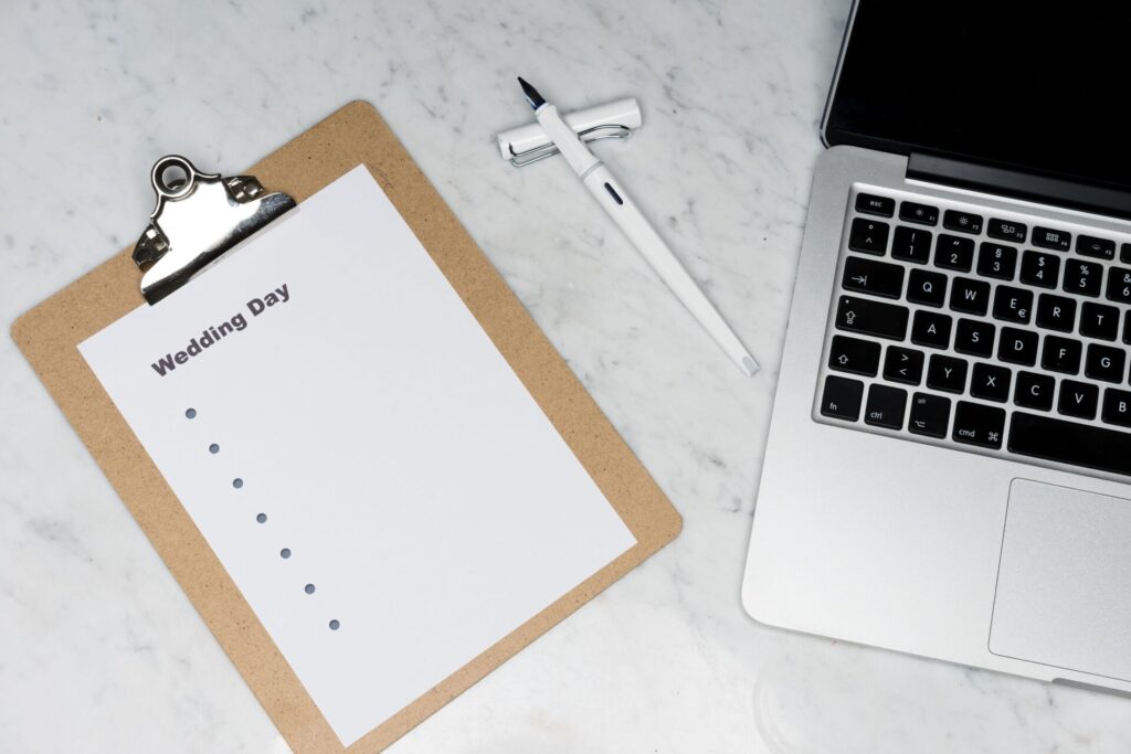 Flat lay of a wedding planning setting with clipboard, laptop, and pen.
