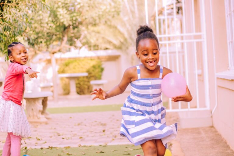 Cheerful cute African American little girls playing together with balloons during party in house yard