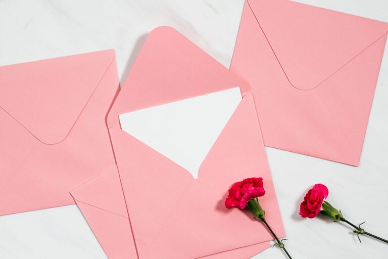 Flat lay of pink envelopes and red carnations on a white marble surface, ideal for invitations.