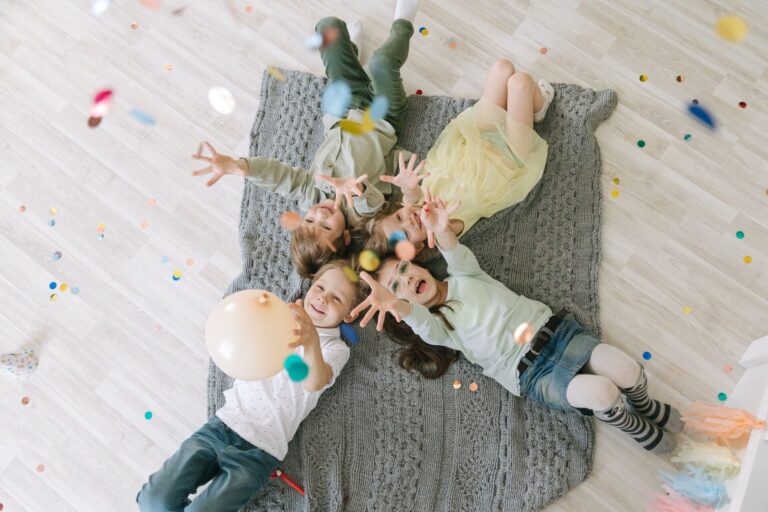 Aerial view of happy children celebrating with balloons and confetti indoors on a rug.
