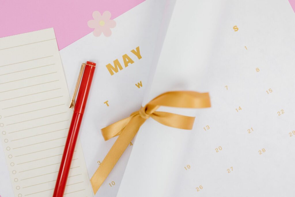 A minimalist flat lay of a calendar with red pen and ribbon on a pink background, perfect for planning.