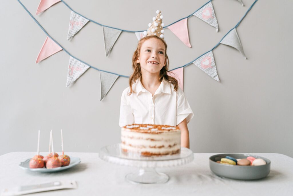 A smiling girl at a birthday party with a cake, wearing a party hat indoors.